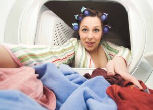 A woman with curlers in her hair reaches into a dryer to pull out a load of laundry.