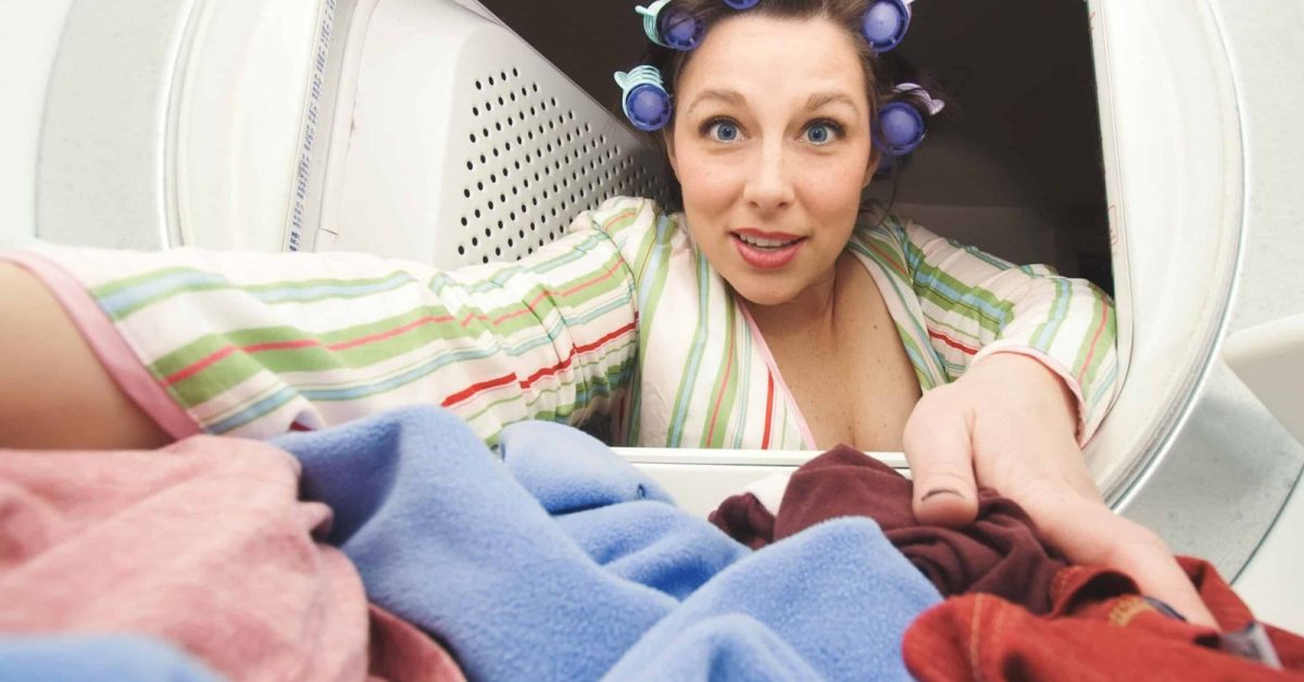 A woman with curlers in her hair reaches into a dryer to pull out a load of laundry.