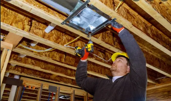 HVAC technician installing metal air duct in unfinished ceiling with power drill during home construction.