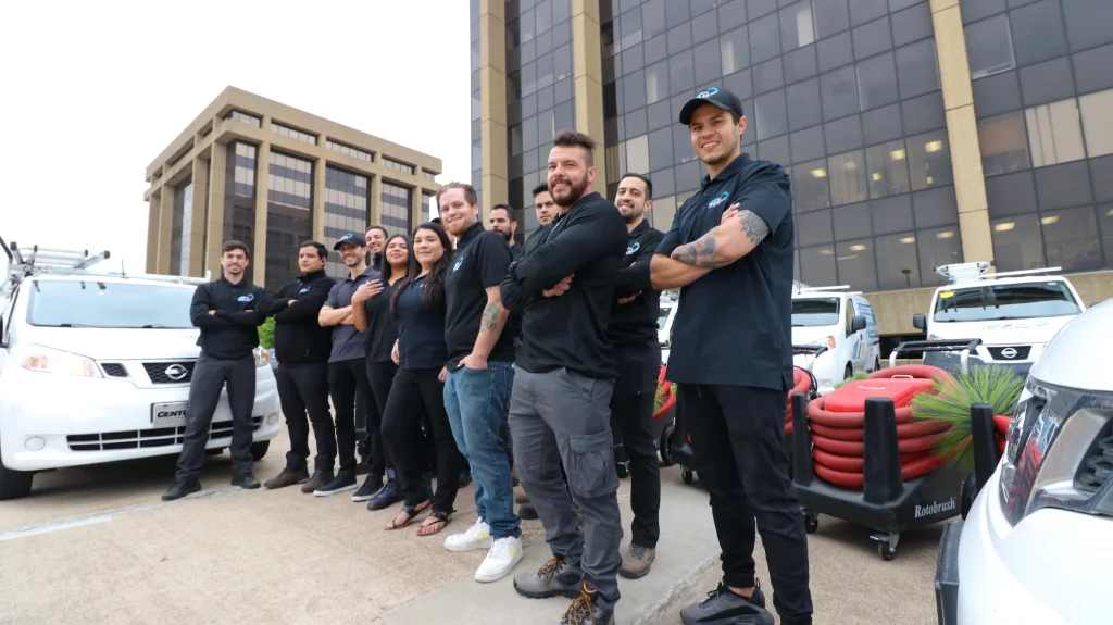 Group of air duct cleaning technicians standing beside service vans and equipment in front of office buildings.