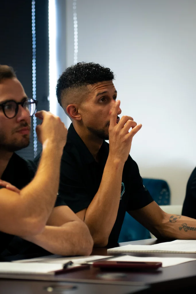 Man attentively listening during a meeting, hand raised to his face while seated beside a colleague with notebooks on the table.